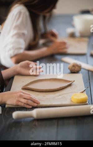 Two women in aprons sitting by table, kneading clay and making earthenware at lesson. Stock Photo