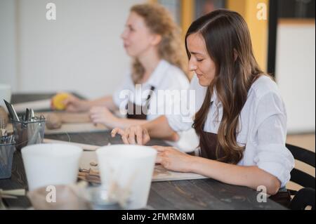 Two women in aprons sitting by table, kneading clay and making earthenware at lesson. Stock Photo