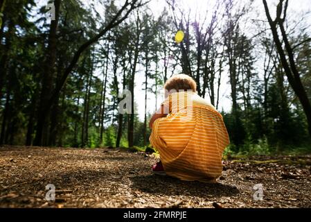 Low to the ground view of the back of a young girl with short ginger hair wearing a yellow dress with white stripes knelt down playing on a woodland f Stock Photo