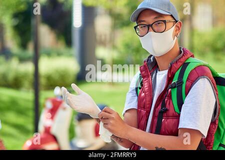 Young asian delivery man wearing face protective mask and gloves looking at camera while standing on a sunny street. Safe delivery during coronavirus Stock Photo