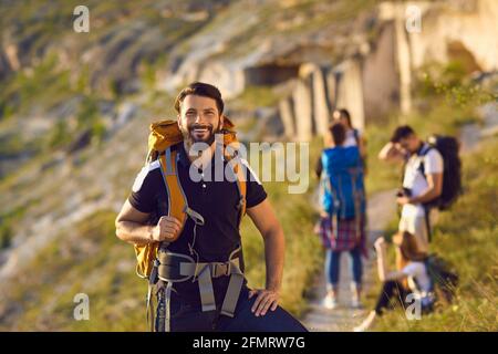 Handsome young man standing on hiking trail and looking at camera with friends in blurred background Stock Photo