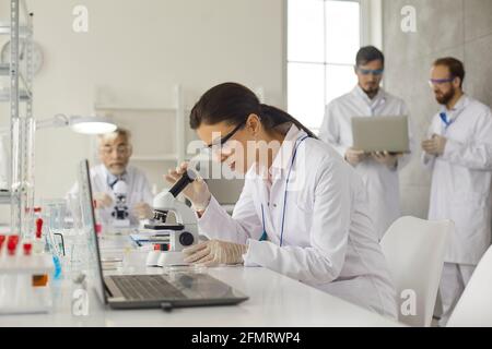 Concentrated female virologist sits at table in laboratory and examines cells under microscope. Stock Photo