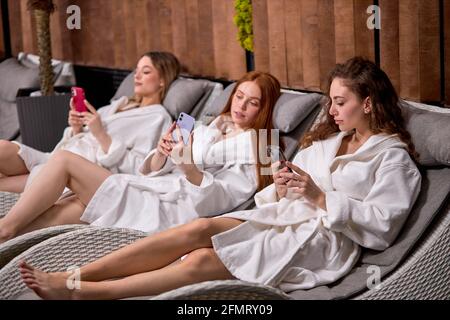 Relaxed girlfriends have rest using smartphones dressed in bathrobes after spa procedures, lying on bed in spa center, enjoying free time. vacation, s Stock Photo
