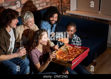 young and happy friends eating pizza and watching movie or tv series at home, american students enjoy spare free time after lessons, having rest after Stock Photo