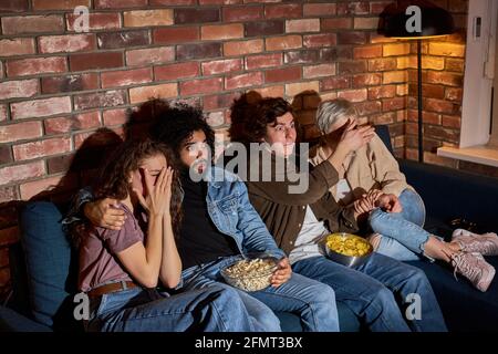 Group of friends watching scary movie on TV at home, eating popcorn, sitting on sofa together, closing eyes, two couples nervous scared in shock terri Stock Photo