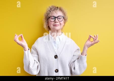 Elderly woman in eyeglasses keep calm, namaste, isolated over yellow background, engaged in yoga meditation, relieve tension. portrait of wrinkled fem Stock Photo