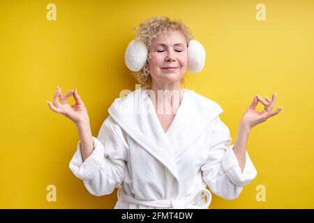Senior woman in bathrobe and headset keep calm, namaste, isolated over yellow background, engaged in yoga meditation, relieve tension. portrait of wri Stock Photo