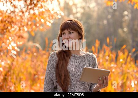 Happy sitting woman using tablet during autumn walks outdoors in park Stock Photo