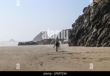 Young man surfer with no shirt and bushy long dark hair walks down misty beach with towering wet rock cliffs toward ocean with waves rolling in - shad