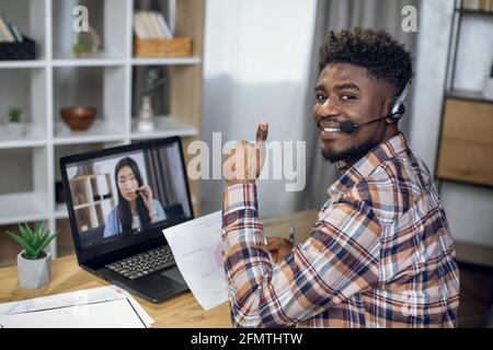 African man smiling and showing thumb up on camera while having video chat on laptop with asian woman. Two coworkers leading briefing remotely from home. Stock Photo