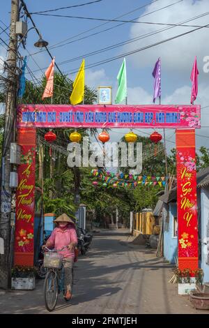 Vietnamese cyclist riding beneath archway decorated with colourful flags and Ho Chi Minh's portrait, Hoi An, Vietnam Stock Photo