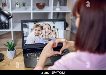 Back view of young woman using wireless laptop for video call with relatives. Pretty female sitting at home and showing with fingers heart shape to express her love. Stock Photo