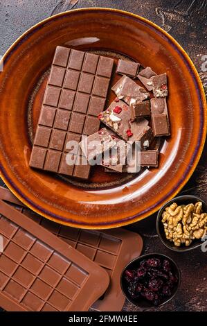 Homemade milk chocolate with hazelnuts, peanuts, cranberries and freeze dried raspberries on a rustic plate. Dark background. Top view Stock Photo