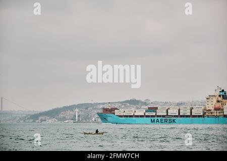 Huge transportation ship  (MAERSK) passing through in istanbul bosphorus during overcast and rainy day with small fishing boat and bridge background. Stock Photo