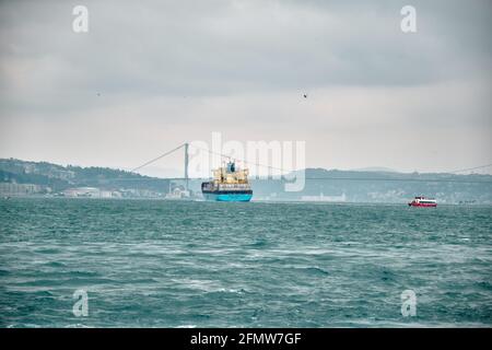 Huge transportation ship passing through in istanbul bosphorus during overcast and rainy day with small fishing boat and bridge background. Stock Photo