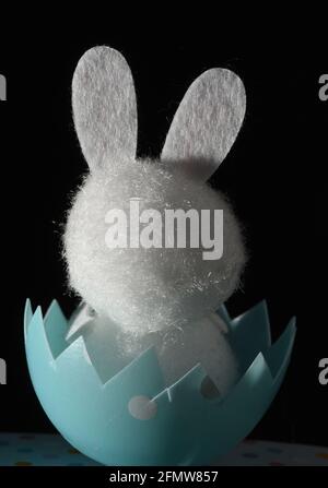 A still life studio vertical image on a black background of the rear view of a white fuzzy toy rabbit sitting in an egg shell, an Easter bunny toy Stock Photo