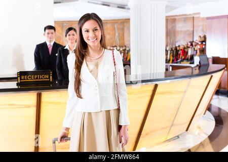 Asian Chinese woman arriving at front desk of luxury hotel in business clothes with trolley Stock Photo