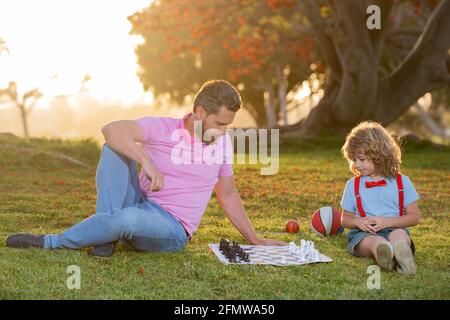 Kid son sitting on grass and playing chess with father. Clever thinking smart child while playing chess. Stock Photo