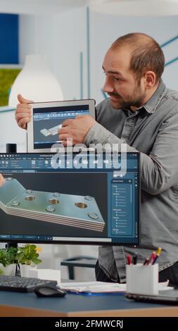 Team of architects and engineers working on building prototype project, using computers running 3D CAD Software. Project manager discussing with employee holding tablet explaining technical specifics Stock Photo