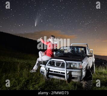 Young woman in the mountains watching beautiful starry night and a comet, sitting on car hood. Illuminated foreground. Bright skyline on background. Astrophotography concept Stock Photo