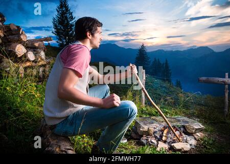 Tyrol - Young man sitting on alpine meadow of a mountain on Campfire in the Bavarian Alps and enjoys the romantic evening sunset of the panorama in le Stock Photo