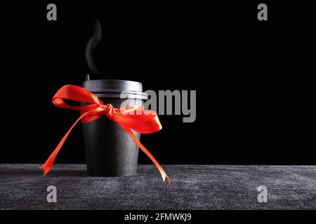 Black paper disposable takeout drink cup with steam over the lid with bow made of gift red ribbon on dark surface and black background Stock Photo