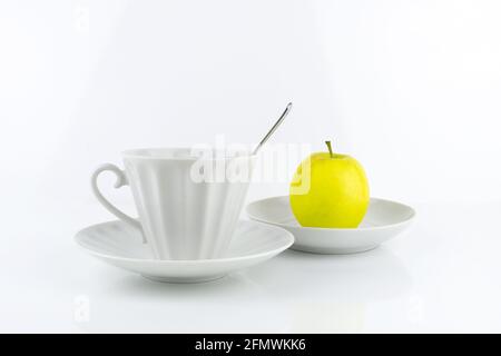 White porcelain cup with spoon and saucer, yellow apple on saucer, on white surface Stock Photo