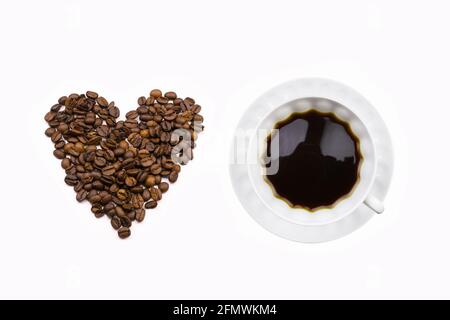Heart made of coffee beans, white cup with coffee and saucer on white background, top view, flatlay. Valentine's Day. Stock Photo