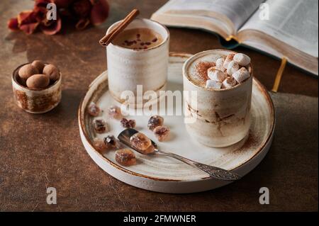 Two cups of coffee with foam and marshmallow and sublimated raspberries with cinnamon on light plate with caramelized sugar and spoon. Nearby is an open book, flowers and candy. Selective focus