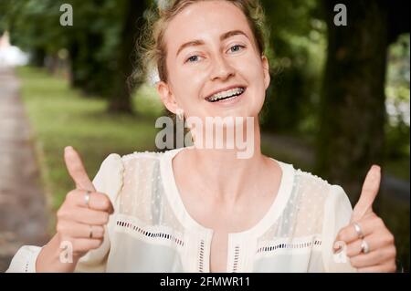 Horizontal image of natural beauty woman outdoors, showing thumbs up, with braces on her teeth smiling. Happy female after dentist visit. Concept of dentistry and oral care Stock Photo