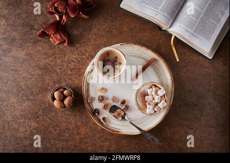 Top view two cups of coffee with foam and marshmallow and sublimated raspberries with cinnamon on light plate with caramelized sugar and spoon. Nearby is an open book, flowers and candy Stock Photo