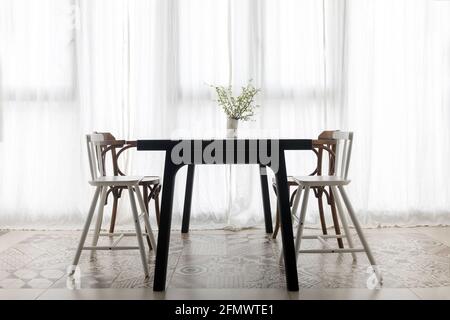 Chairs placed near dining table with bouquet of flowers in vase against window with white curtain in modern minimalistic style apartment Stock Photo