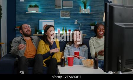 Multiracial friends laughing smiling having fun together while sitting on couch in front of TV watching comedy movie. Group of mixed race people enjoying free time drinking beer eating snacks Stock Photo