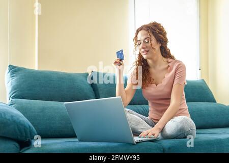 Smiling millennial girl sit on couch using laptop buying on Internet enter credit card details, easy banking system service concept. High quality photo Stock Photo