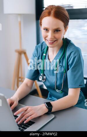 Close-up face of attractive smiling young female doctor in blue green medical uniform sitting at desk with laptop on background of window Stock Photo