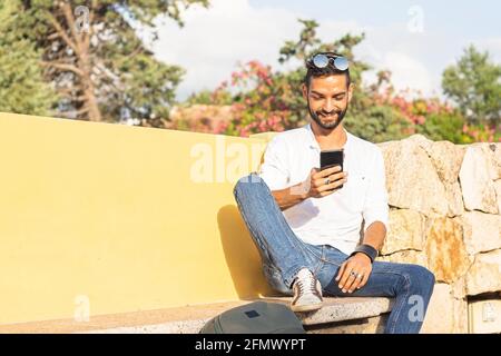 Young beautiful happy stylish guy with backpack sitting on a city bench waiting for a public transportation way to his travel destination using smartp Stock Photo