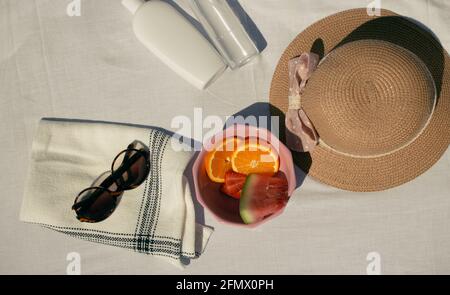 Horizontal image of fruits (orange,watermelon, strawberries )in a pink bowl together with summer items on a white fabric background. Summer 2021 Stock Photo