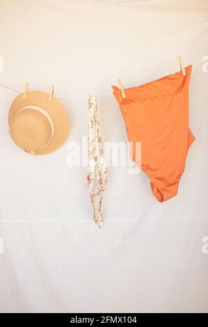 Vertical image of a summer hat, a belt and an orange swimsuit on a clothesline on a white isolated background. Summer Concept 2021. Stock Photo