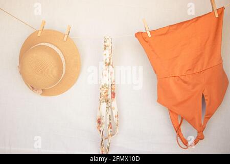 Horizontal image of a summer hat, a belt and an orange swimsuit on a clothesline on a white isolated background. Summer Concept 2021. Stock Photo