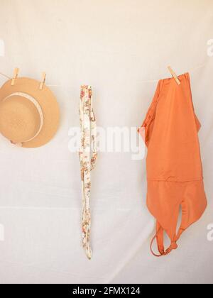 Vertical image of a hat, a belt and an orange swimming costume on a clothesline on a white isolated background. Summer Concept 2021. Stock Photo