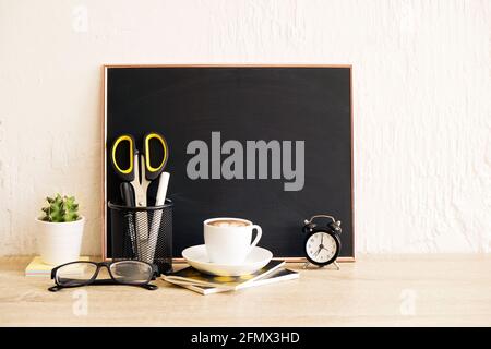 Blank chalk board, scissors, folded glasse, green cactus plant, white cup of cappuccino coffe beverage, mini alarm clock on wooden table. Background,