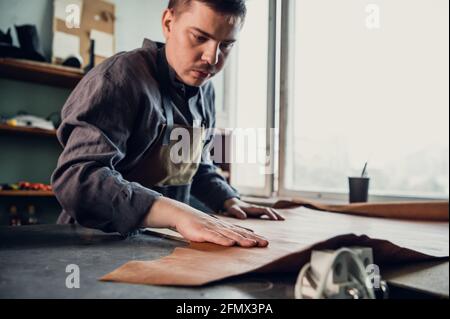 A young apprentice in a boot workshop prepares leather for further use on a large table. Stock Photo