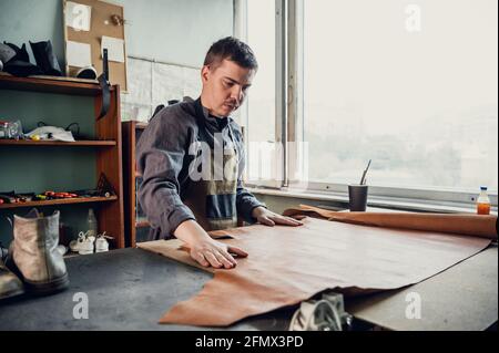 A young apprentice in a boot workshop prepares leather for further use on a large table. Stock Photo