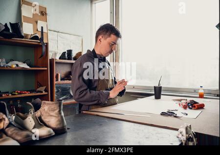 A young shoemaker makes a drawing for a pattern for leather shoes on a table in his workshop. Stock Photo