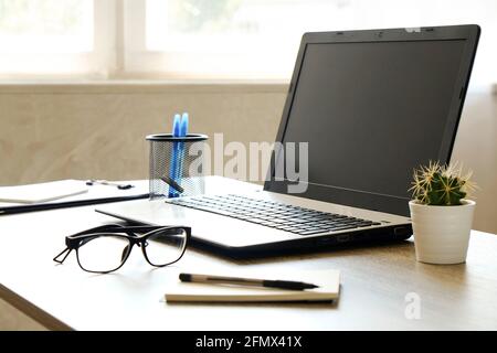 Laptop computer, clipboard, cactus home plant, supplies and folded glasses on wooden desk in spacious office full of sunlight. Designer's creative wor Stock Photo