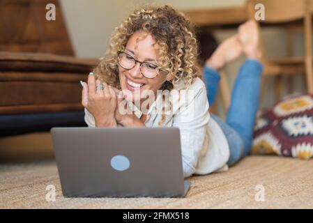 Cheerful pretty adult woman do video call online with laptop laying on the floor at home - relax and enjoy laptop and internet connection people - mod Stock Photo