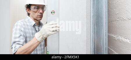 Man drywall worker or plasterer checking level of white plasterboard wall with bubble level at construction site. Wearing white hardhat, work gloves a Stock Photo