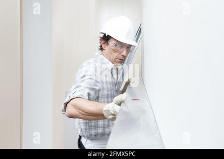 Man drywall worker or plasterer checking level of white plasterboard wall with bubble level at construction site. Wearing white hardhat, work gloves a Stock Photo