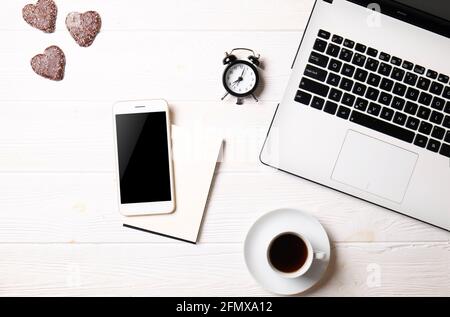 Flat lay on white wooden workspace with laptop computer, cell phone gadget, supplies, mini alarm clock & black coffee. Wood textured office desk. Dead Stock Photo