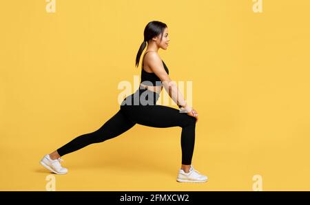 African american lady in sportswear stretching legs before workout, preparing for sport training on yellow background Stock Photo